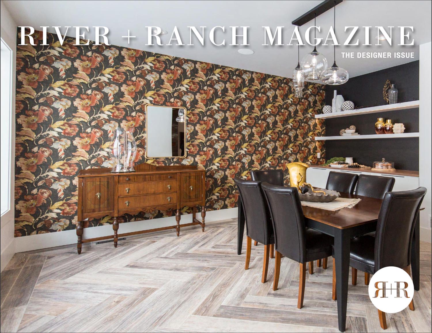 Dining room with dark floral wallpaper, a wooden sideboard, and a dark dining table with black leather chairs. Modern glass pendant lights hang above styled shelves. RIVER + RANCH MAGAZINE and a Press interior design services North Dakota feature are atop the table.