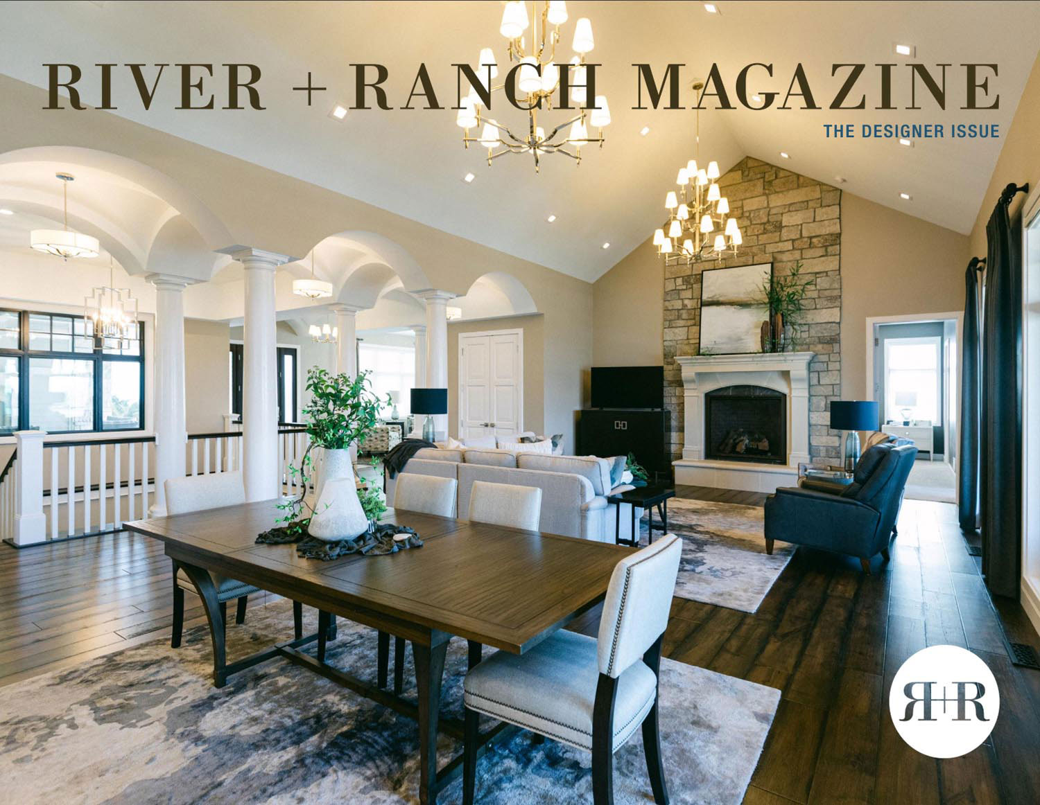 Spacious, elegant living and dining area with vaulted ceilings, chandeliers, stone fireplace, and modern furnishings—showcasing Press interior design services North Dakota—featured on the cover of River + Ranch Magazine, “The Designer Issue.”.
