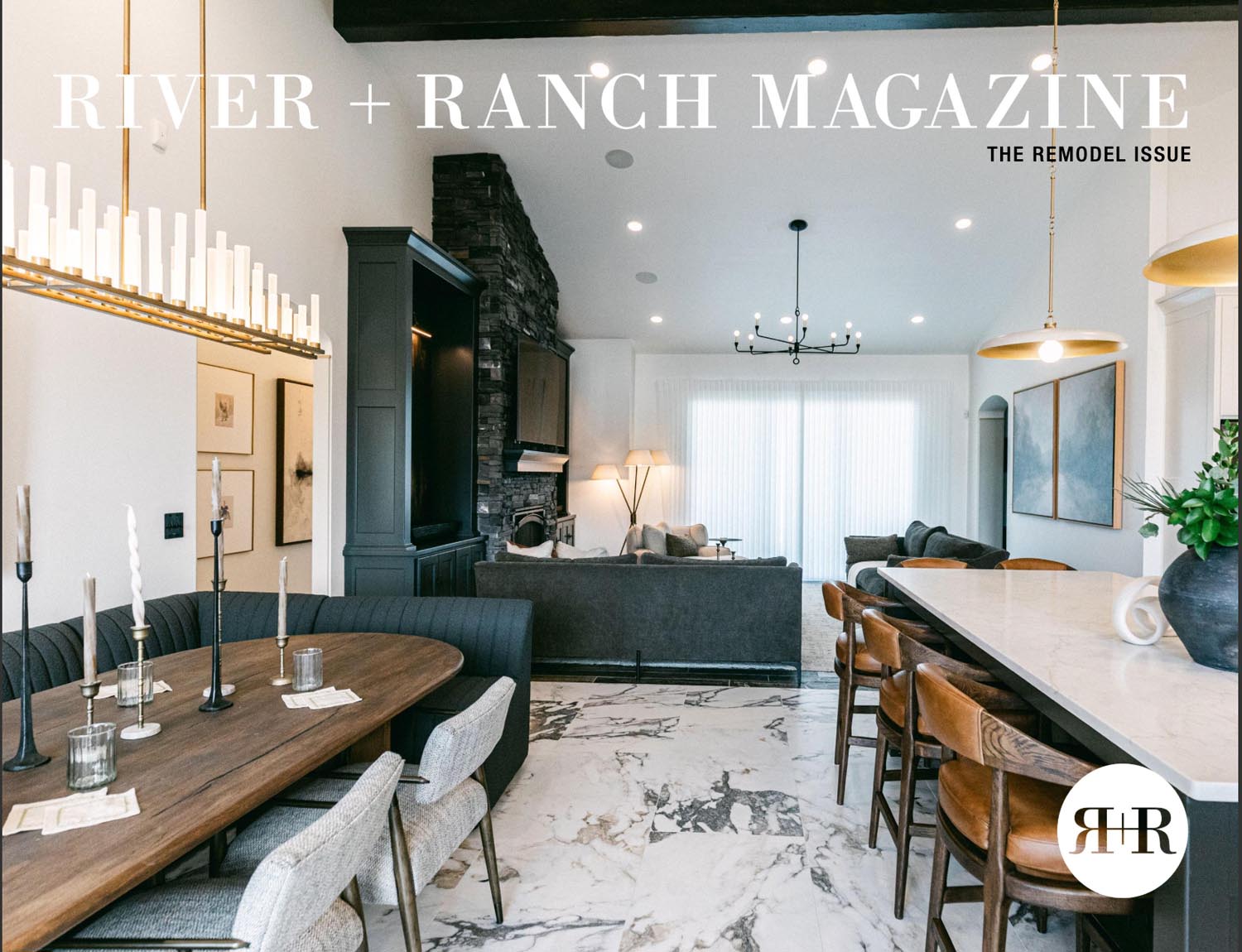 A modern open-concept living and dining area with a marble floor, dark wood dining table, gray chairs, stone fireplace, and large kitchen island under elegant chandeliers. Text reads “RIVER + RANCH MAGAZINE: THE REMODEL ISSUE.”.